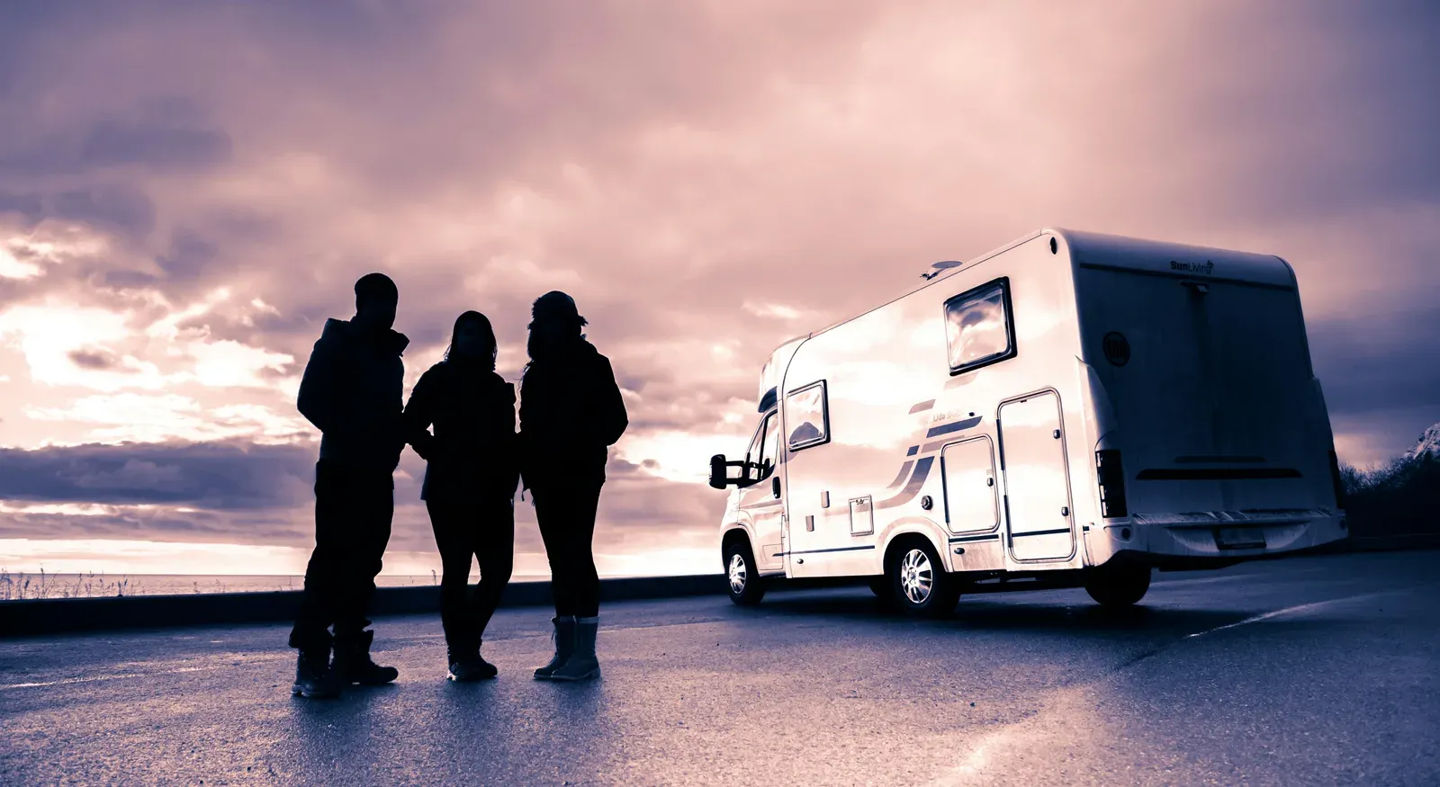 Silhouettes of a family standing beside a motorhome on a coastal road at sunset
